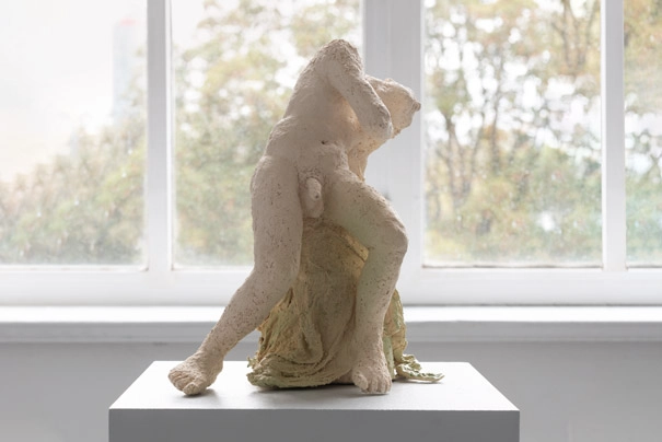 Gezeigt wird eine durch Pigmente glasierte figurative Keramik aus weißem Ton auf einem Sockel vor einem Fenster.